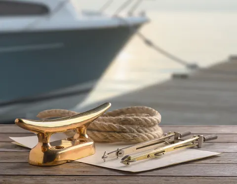 A polished brass cleat, a coiled rope, and a pair of brass dividers on a weathered dock, with a moored boat out of focus behind them