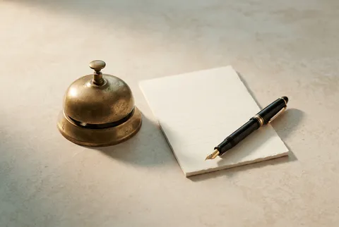 A brass desk bell beside a cream notepad and a fountain pen on a warm stone surface
