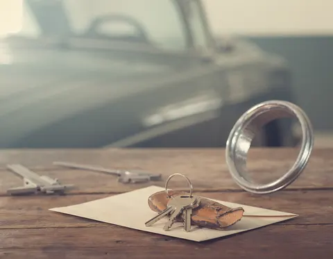 Car keys on a worn leather fob, a chrome headlight bezel, and a steel caliper on a wooden workbench, with a classic car out of focus behind them