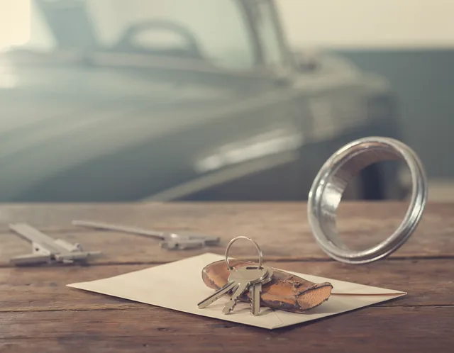 Car keys on a worn leather fob, a chrome headlight bezel, and a steel caliper on a wooden workbench, with a classic car out of focus behind them